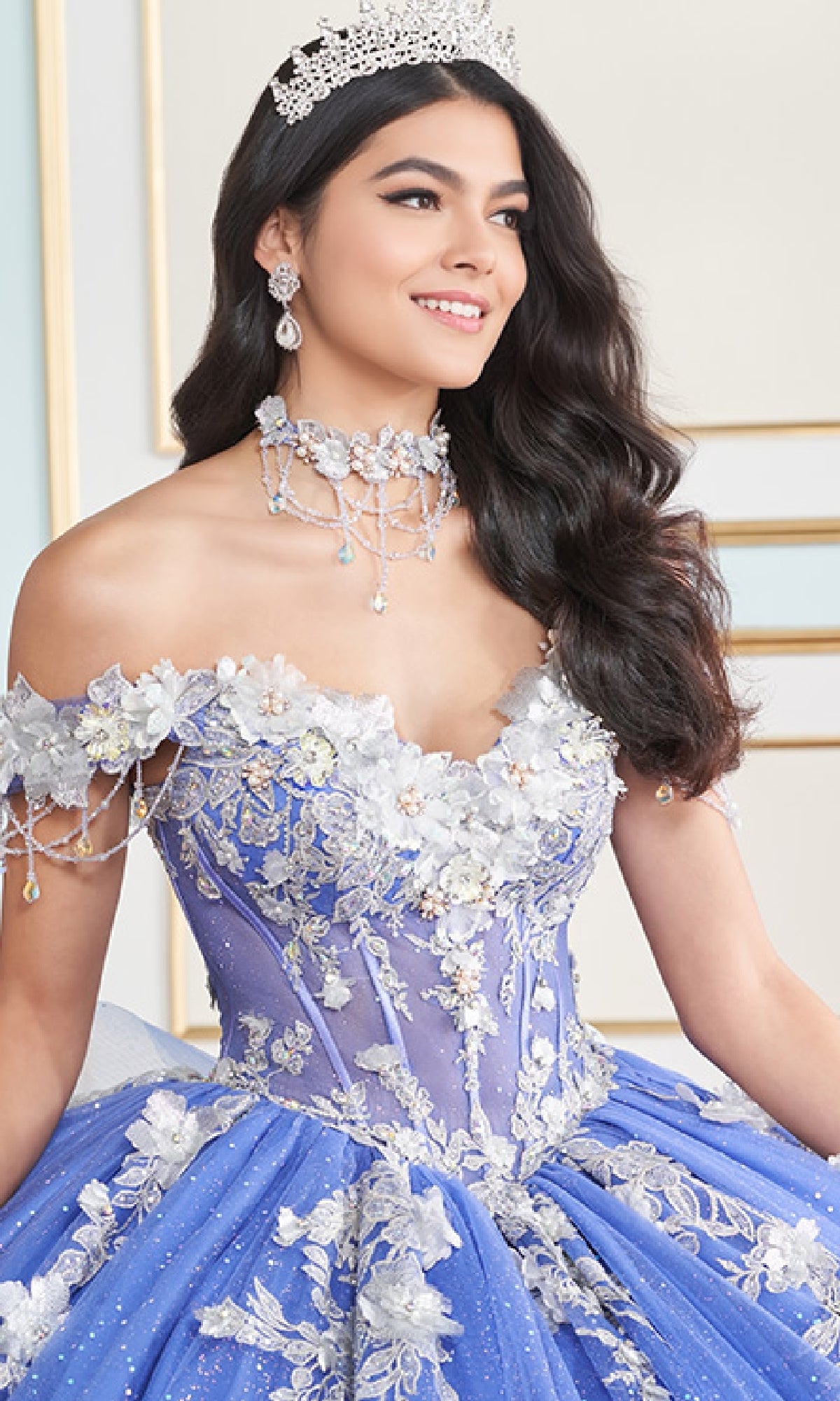 A woman wearing the Princesa Quinceanera Dress PR30178—a blue, off-the-shoulder gown with a corset bodice, floral appliqués, and beadwork—smiles indoors with a jeweled tiara.